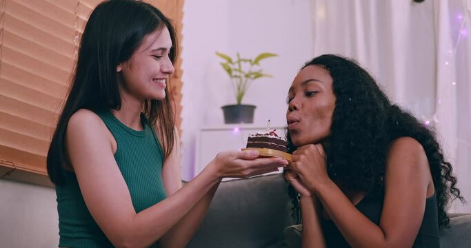 Young woman surprising her girlfriend with cake at home. Female lesbian couple celebrating birthday in living room.