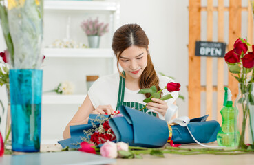 Asian professional successful female florist designer shop owner entrepreneur businesswoman sitting smiling holding decorating picking choosing red roses flowers bouquet in floral garden store studio