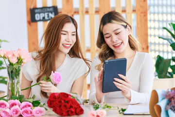 Asian professional successful female florist designer flower shop owner entrepreneur decorating pink roses bouquet in glass vase when colleague checking stock online via tablet in floral store studio