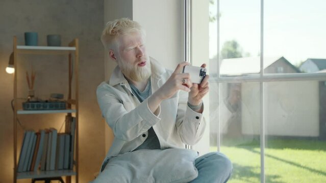 Albino man with white beard making photos of outdoor view sitting at the window. Young man makes several shots, satisfied with result. Carefree lifestyle. High quality 4k footage