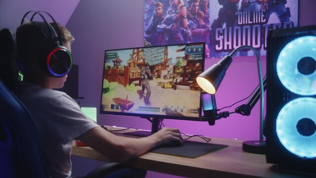 Young gamer plays third person shooter on personal computer. Schoolboy in headphones enjoys online video game at home. Desk illuminated by RGB LED strip light. Concept of gaming at home. Zoom in.