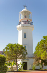 A white lighthouse surrounded by green plants against a blue sky on a sunny morning