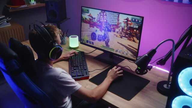 Young gamer plays third person shooter on personal computer. Schoolboy in headphones enjoys online video game at home. Desk illuminated by RGB LED strip light. Concept of gaming at home. Dolly shot.