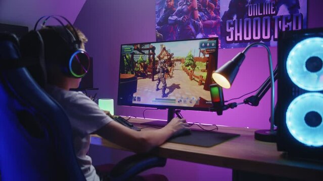 Young gamer plays third person shooter on personal computer. Schoolboy in headphones enjoys online video game at home. Desk illuminated by RGB LED strip light. Concept of gaming at home. Back view.