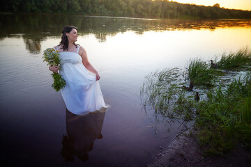 Slavic plump plump chubby girl in long white dress on the feast of Ivan Kupala with flowers and water in a river or lake on a summer evening