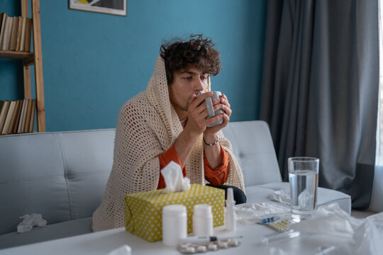 Cold medicine, sick man drinking hot beverage to get well from flu and fever. Dirty paper tissues and pills on table. Ill person wearing warm woolen plaid.