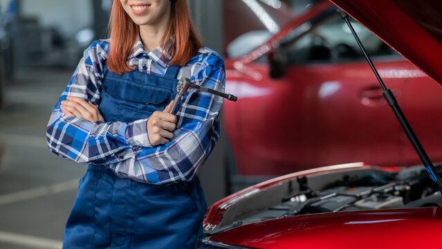 A woman auto mechanic stands at the open hood of a car and holds a screwdriver. 