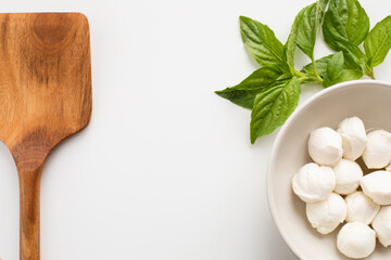 Closeup top view on a bowl with fresh mozzarella, basil leaves and wooden spatula on white background with copy space