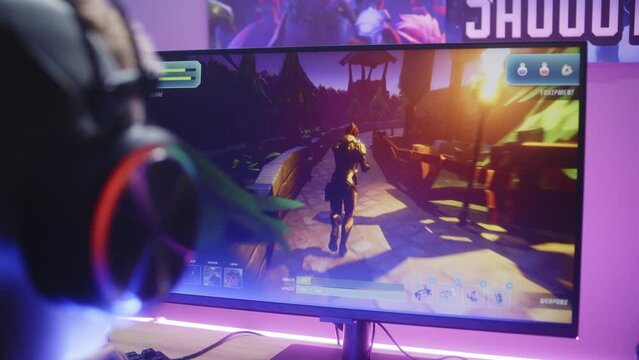 Young boy in headphones plays third person shooter. Gamer enjoys online video game on home personal computer. Desk illuminated by RGB LED strip light. Concept of gaming at home. Back view. Close up.