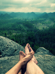 Crossed bare legs of a heterosexual couple cuddling on a cliff. Park background with rocks and forests.