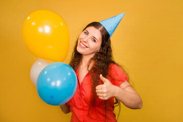Fototapeta na wymiar Portrait of a young woman in a festive cap with multi-colored balloons in her hand isolated on a yellow background. Noisy birthday concept.