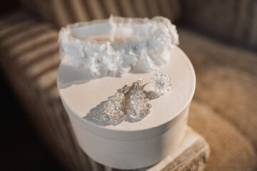 A white wedding leg band and elegant crystal earrings stand on a white round basket. Gift box. Wedding details