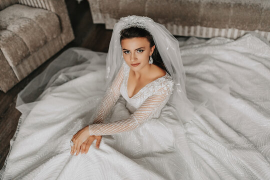 A brunette bride wearing a lace dress poses while sitting on the floor. Beautiful hair and make-up, open bust. Wedding portrait. Gorgeous silver jewelry