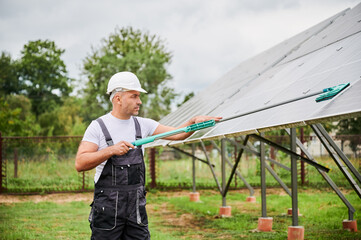 Male adult carefully wiping dust from PV solar panel. Professional worker taking care about battery cleanliness. Worker in helmet and overalls cleaning solar batteries.