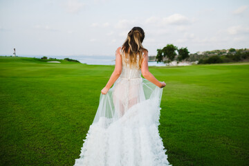 Fototapeta premium A beautiful, young girl with blond hair runs across the lawn in the light of the setting sun, in a white, stylish wedding dress, rear view.
