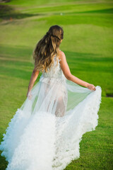 Fototapeta na wymiar A beautiful, young girl with blond hair runs across the lawn in the light of the setting sun, in a white, stylish wedding dress, rear view.