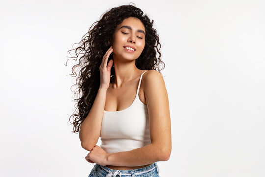 beautiful woman in a white T-shirt and blue jeans with long curly hair stands against a white background and smiles with her eyes closed in pleasure