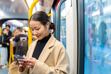 Asian woman using mobile phone and listening to the music on earphones during travel on train in the city. Attractive girl enjoy urban lifestyle in the city with using wireless technology on device.