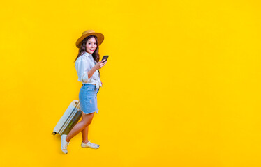 Fototapeta na wymiar Pretty Asian woman passenger in trendy fashion is using mobile phone while carrying her luggage bag in happiness for travel and summer vacation isolated on yellow background