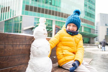 a happy smiling boy made a little snowman on a bench in the center of a metropolis, a winter city