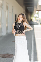 Obraz premium Fashion beautiful young girl in a stylish black lace top with a bra and a white skirt with a handbag walks on the street. Hipster woman with tattoos on her arms