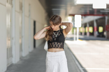 Stylish beautiful young woman in a fashionable black lace top with a bra and a white skirt with a handbag walks in the city and straightens her hair