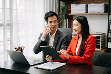 Two Asian business workers talking on the smartphone and using laptop while sitting in office room.