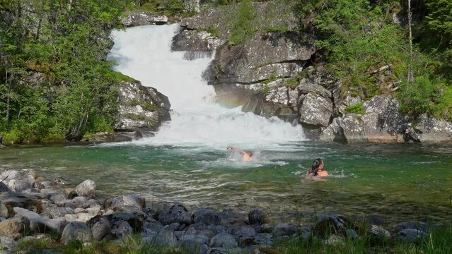 Couple going for a swim in ice cold fresh Norwegian mountain river - Static summer clip with man and woman going in and out of water - Beautiful waterfall background