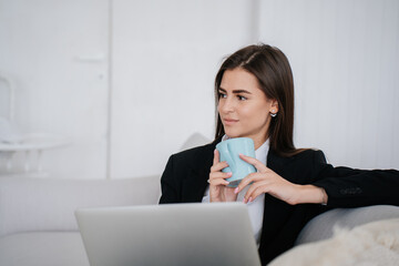 Young female entrepreneur in black suit sitting on couch with laptop holds cup of coffee looks away with pensive face expression at home.Purposeful American businesswoman works home.