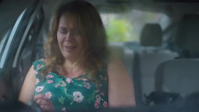 Depressed Caucasian woman crying sitting in car and screaming. Shooting through windshield of frustrated angry plus-size lady shouting in automobile outdoors