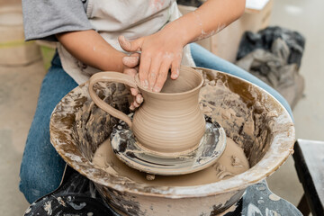 Cropped view of young female artist in apron creating clay jug on pottery wheel on table while working in ceramic workshop, artisanal pottery production and process