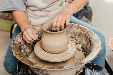 Cropped view of brunette female artisan in apron cutting clay vase on pottery wheel while working in ceramic workshop at background, clay shaping and forming process