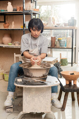 Young asian female potter in apron molding wet clay on pottery wheel near tools and bowl with sponge while working in ceramic studio at background, clay sculpting process concept
