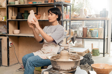 Young asian female artist in apron holding ceramic sculpture while sitting near clay on pottery wheel and tools in blurred studio at sunset, clay sculpting process concept