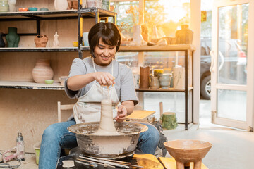 Smiling young asian female artist in apron pouring water on clay on pottery wheel near tools and...