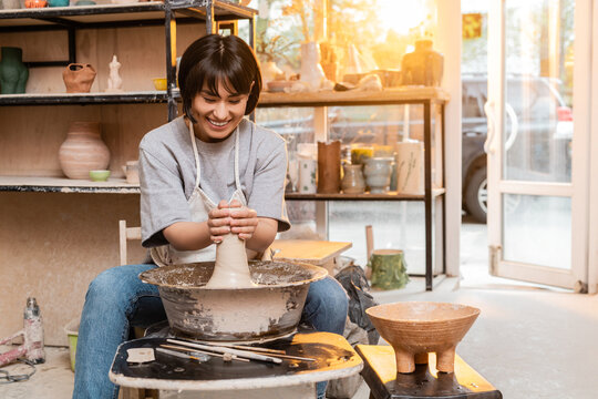 Cheerful young asian female potter in apron molding clay on pottery wheel near bowl and tools in ceramic studio at background at sunset, artisan creating unique pottery pieces - Powered by Adobe