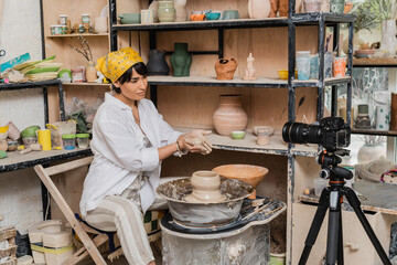 Young asian female artisan in workwear and headscarf holding wet clay near digital camera on tripod and pottery wheel in ceramic workshop, clay sculpting process concept