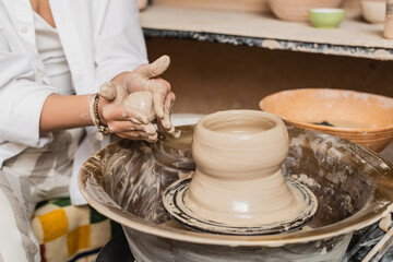 Cropped view of young female potter in workwear holding wet clay near pottery wheel and blurred bowl in blurred ceramic studio, clay sculpting process concept