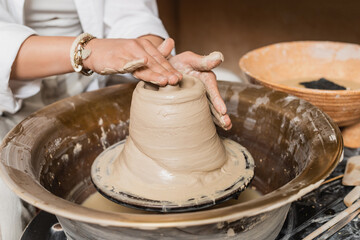 Cropped view of young female artist molding wet clay on pottery wheel while working near blurred bowl with water and tools in workshop, pottery studio workspace and craft concept