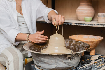 Cropped view of blurred female artist in workwear pouring water on clay on pottery wheel near tools and bowl in ceramic workshop at background, pottery artist showcasing craft