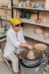 Young asian brunette artisan in headscarf and workwear pouring water on clay on pottery wheel near...