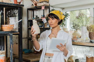 Young brunette asian female artisan in headscarf and workwear holding clay sculpture and digital tablet while working in ceramic workshop, creative process of pottery making