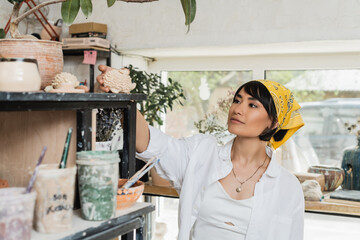 Young female asian artist in workwear and headscarf taking clay product from shelf while standing near pottery tools in pottery class, pottery studio with artisan at work