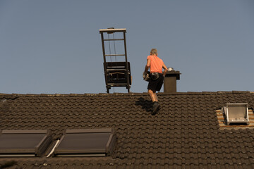 Roofer with an orange t-shirt walks on the roof with gray roof tiles and carries a stack of old...