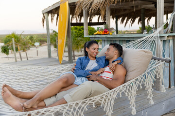 Biracial romantic young couple looking at each other and lying on hammock at beach