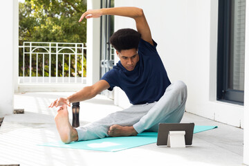 Biracial man doing yoga, stretching and using tablet sitting on sunny balcony