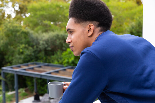 Happy, thoughtful biracial man standing on balcony, looking into garden holding cup of coffee