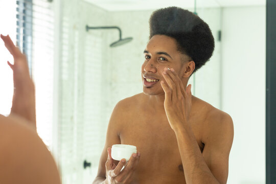 Happy biracial man looking in mirror and applying face cream in sunny bathroom - Powered by Adobe