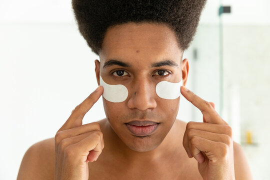 Biracial man looking in mirror and applying under eye masks in sunny bathroom