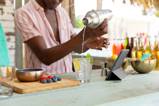 African american male bartender preparing cocktails using shaker at beach bar, unaltered